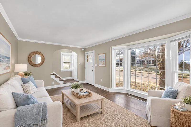 After: the same living room virtually staged in coastal style with a white linen sofa, blue throw pillows, a light wood coffee table on a jute rug, a slipcovered armchair by the bay window, a round rattan mirror, and ocean artwork